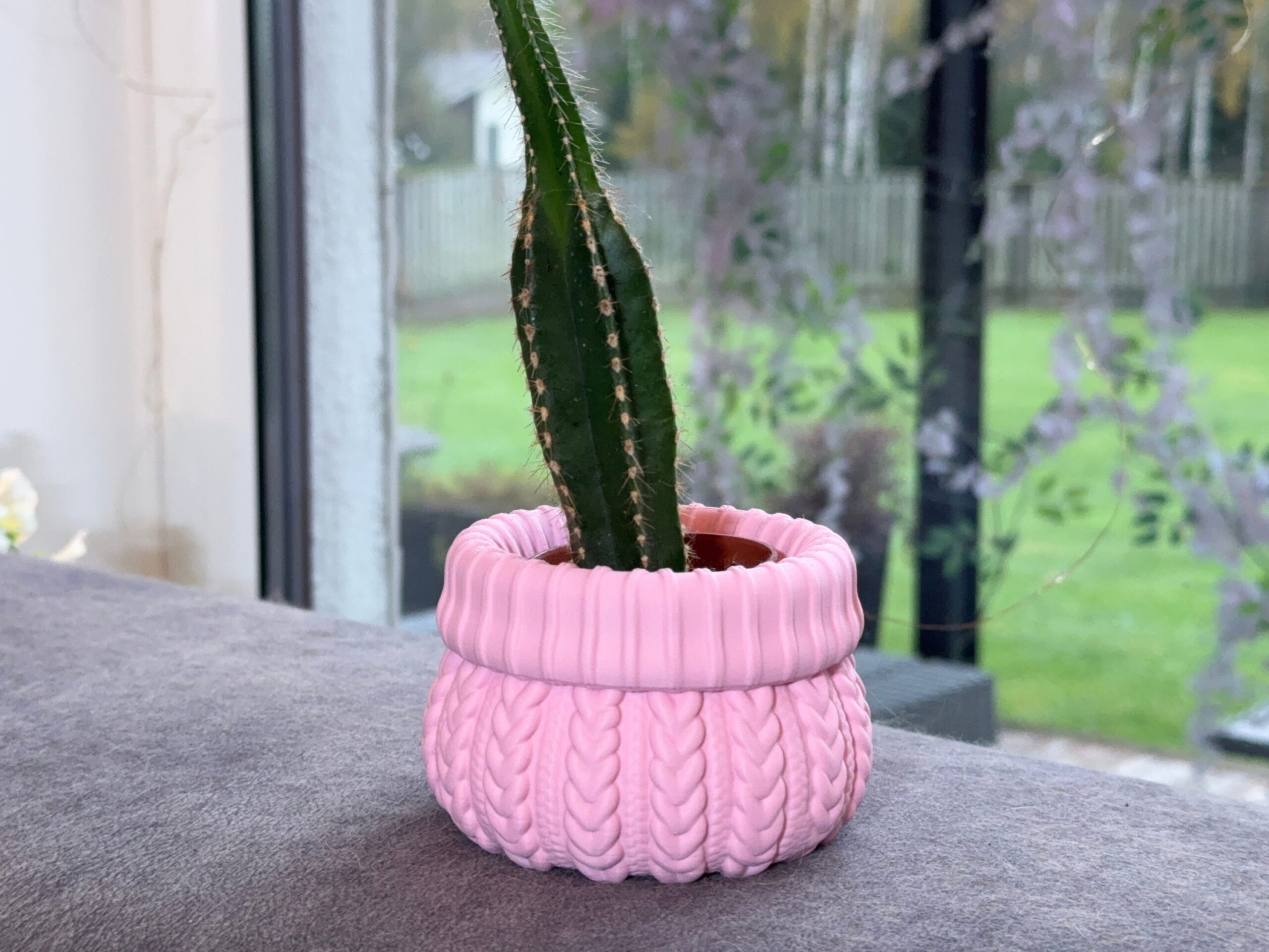 Pink cable knit planter holding a tall cactus in a separate nursery pot beside a window.